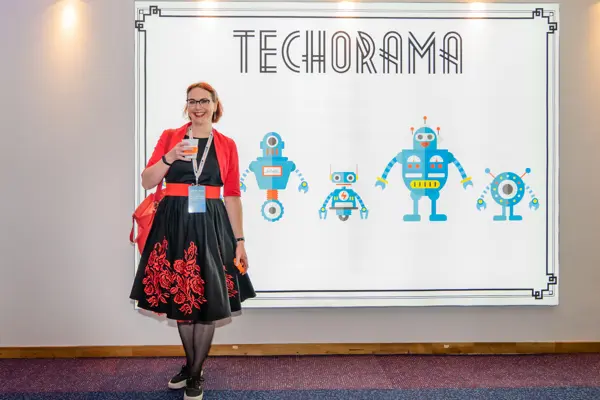 Stacy in front of techorama poster in a black dress holding a coffee